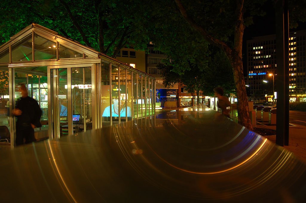 The glass pavilion at Worringer Platz, Düsseldorf, at night, the streamed face glowing inside