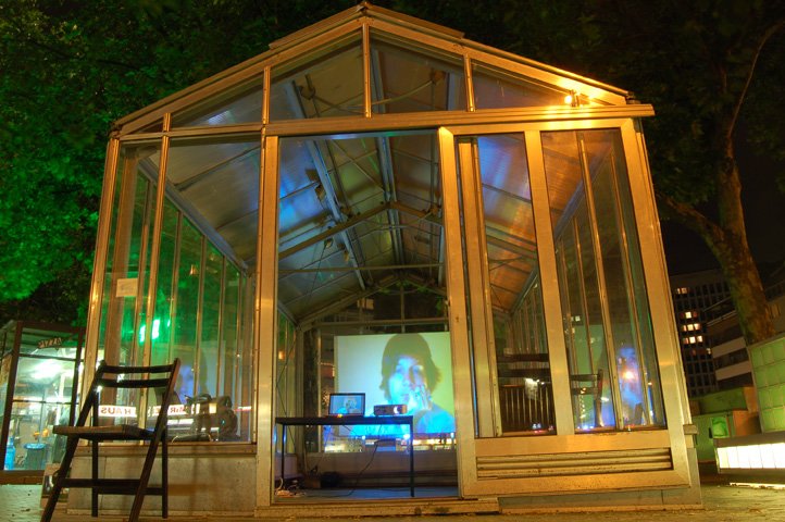 A glass pavilion lit at night, the artist's live webcam face projected inside it